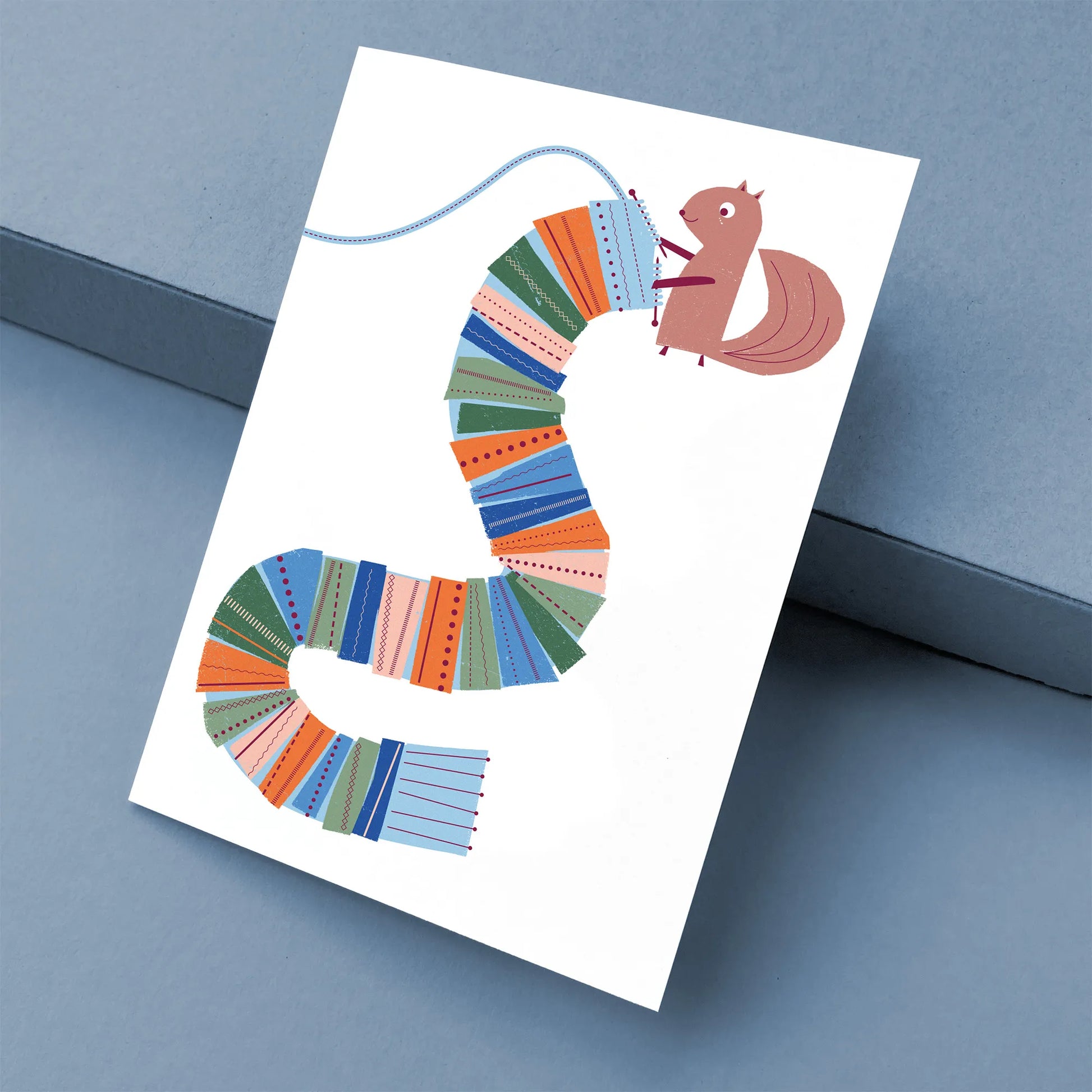 Kautzi greeting card featuring a squirrel knitting a long, colourful scarf on a white background, shown at an angle on a blue surface with a kraft envelope – designed in Cape Breton, Nova Scotia.