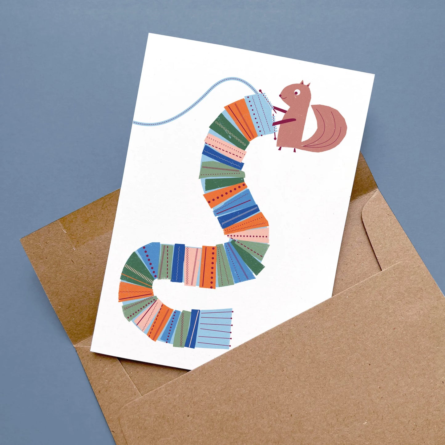 Kautzi knitting squirrel greeting card with a squirrel knitting a long, striped scarf in bright colours, displayed flat with a kraft envelope on a blue background – designed in Cape Breton, Nova Scotia.