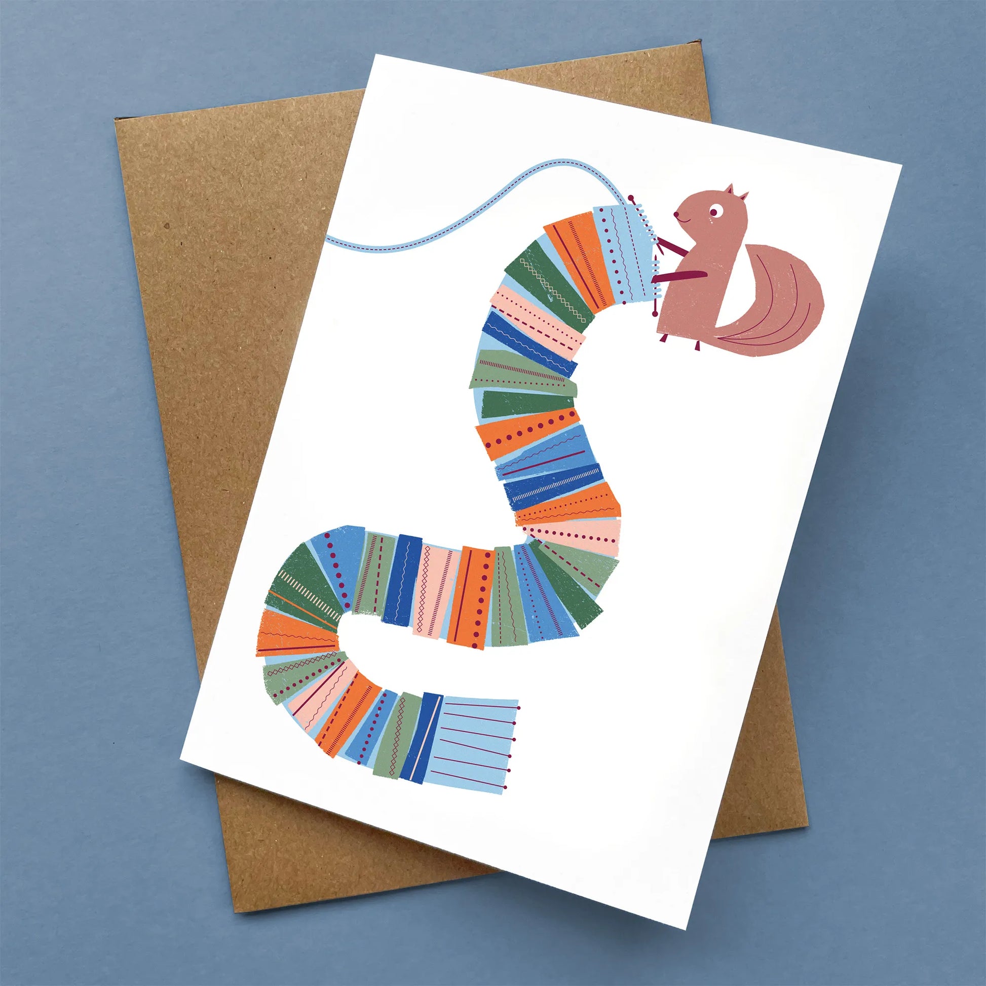 Kautzi greeting card showing a squirrel knitting a long, patchwork-style scarf in shades of blue, green, orange, and pink, paired with a kraft envelope – designed in Cape Breton, Nova Scotia.