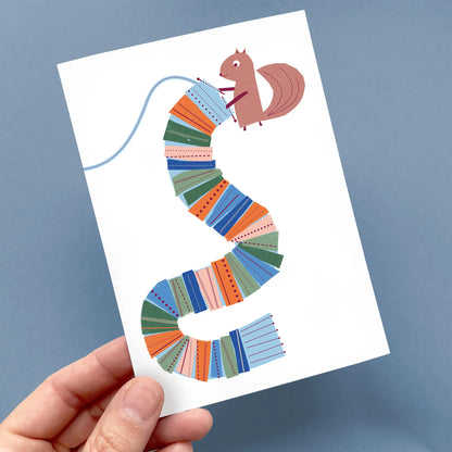 Hand holding a Kautzi greeting card featuring a squirrel knitting a colourful scarf with playful patterns – designed in Cape Breton, Nova Scotia.
