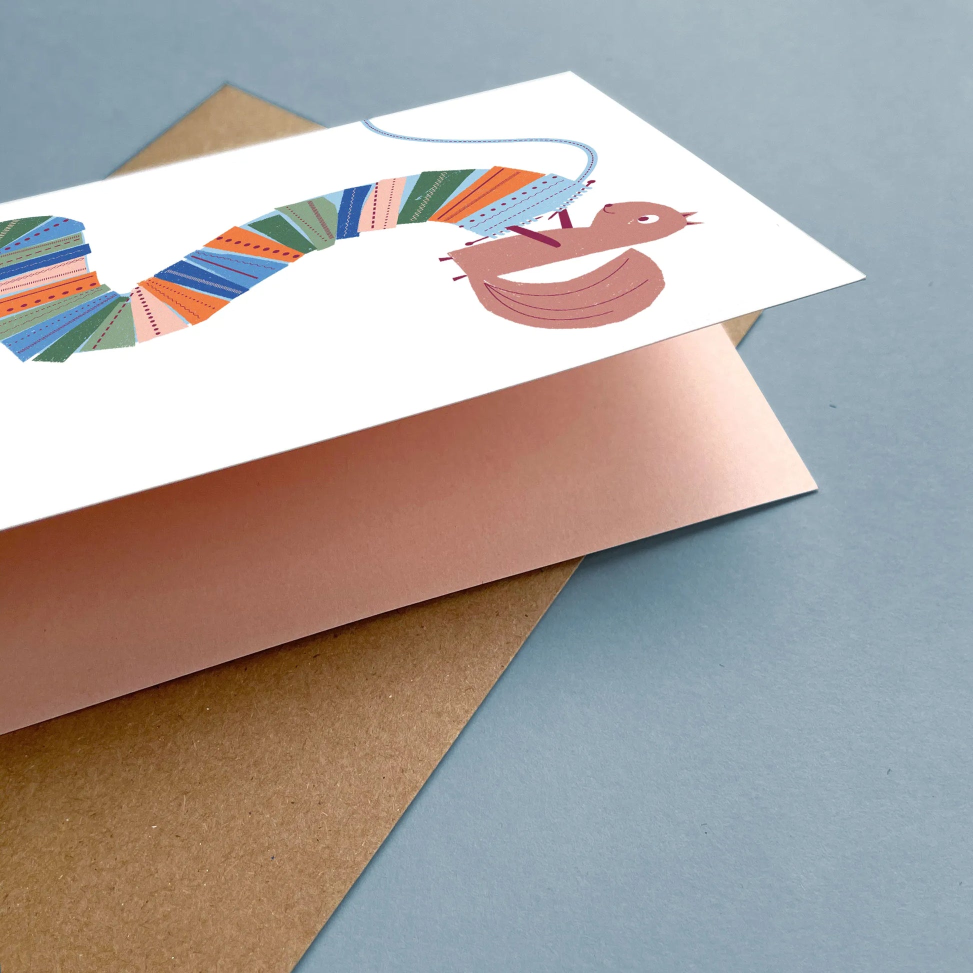 Kautzi knitting squirrel greeting card partially open to show the printed pink interior, featuring a squirrel knitting a long, colourful scarf, with a kraft envelope on a blue background – designed in Cape Breton, Nova Scotia.