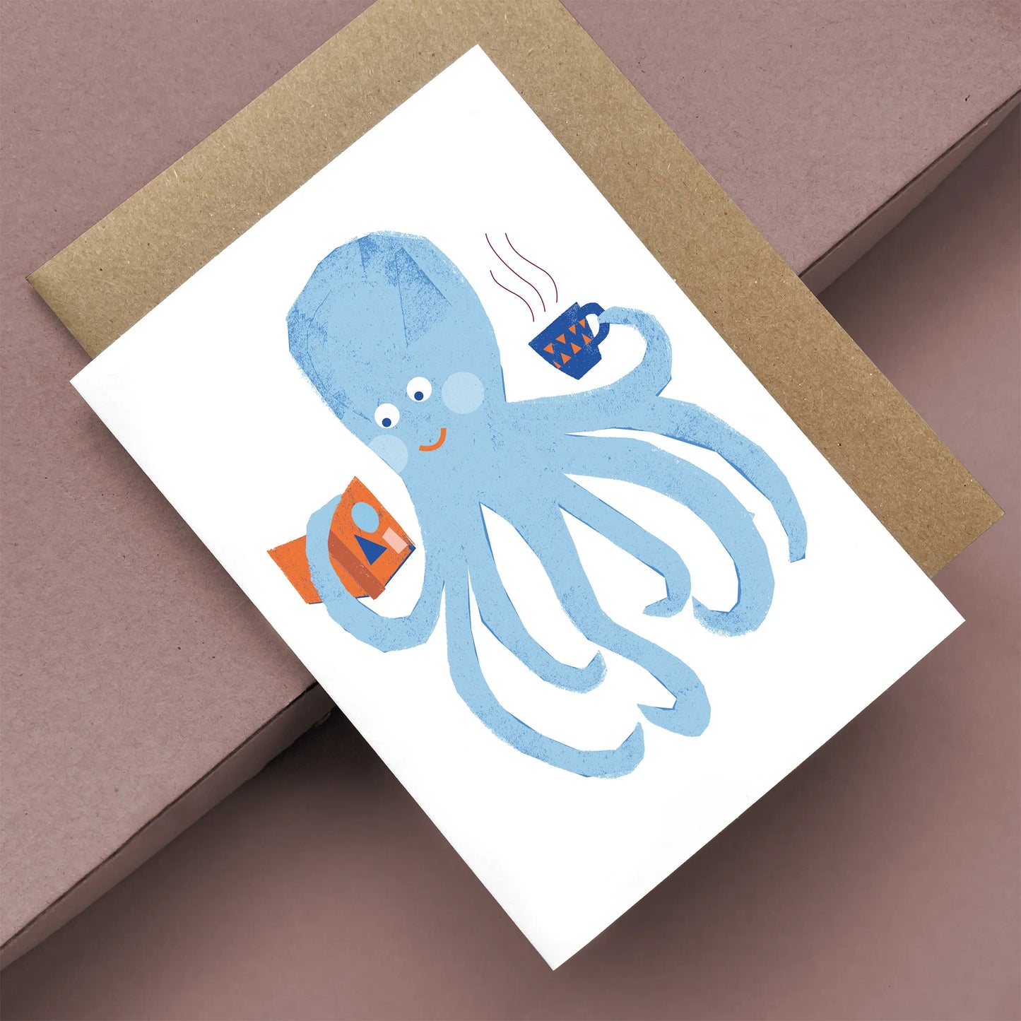 Kautzi greeting card featuring a cheerful blue octopus holding a steaming cup and a book, shown at an angle on a mauve background with a kraft envelope – designed in Cape Breton, Nova Scotia.