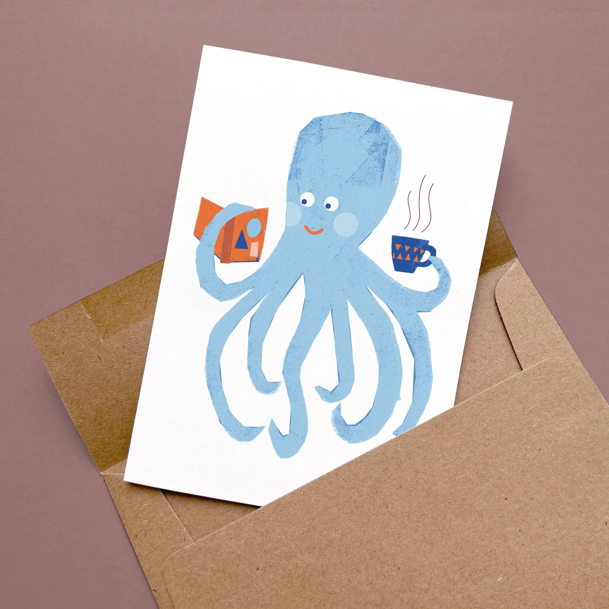 Kautzi octopus greeting card with a blue octopus reading a book and holding a cup of coffee, displayed flat with a kraft envelope on a mauve background – designed in Cape Breton, Nova Scotia.