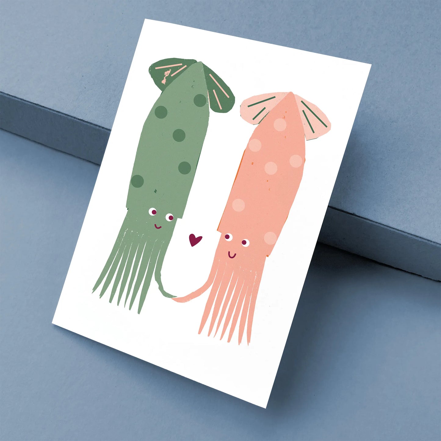 Kautzi greeting card featuring two squids in green and coral pink with a small heart between them, shown at an angle on a blue background – designed in Cape Breton, Nova Scotia.