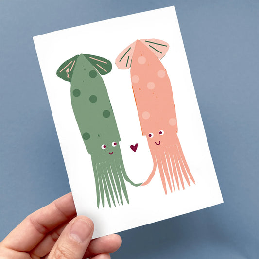 Hand holding a Kautzi greeting card featuring two friendly squids in green and coral pink with a heart between them – designed in Cape Breton, Nova Scotia.