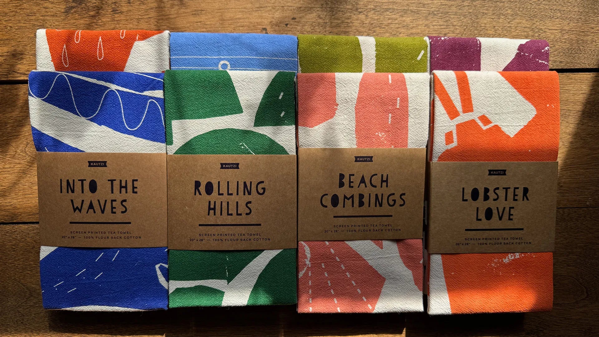 Eight Kautzi screen-printed flour sack tea towels arranged on a wood surface, showing the full collection of East Coast-inspired designs