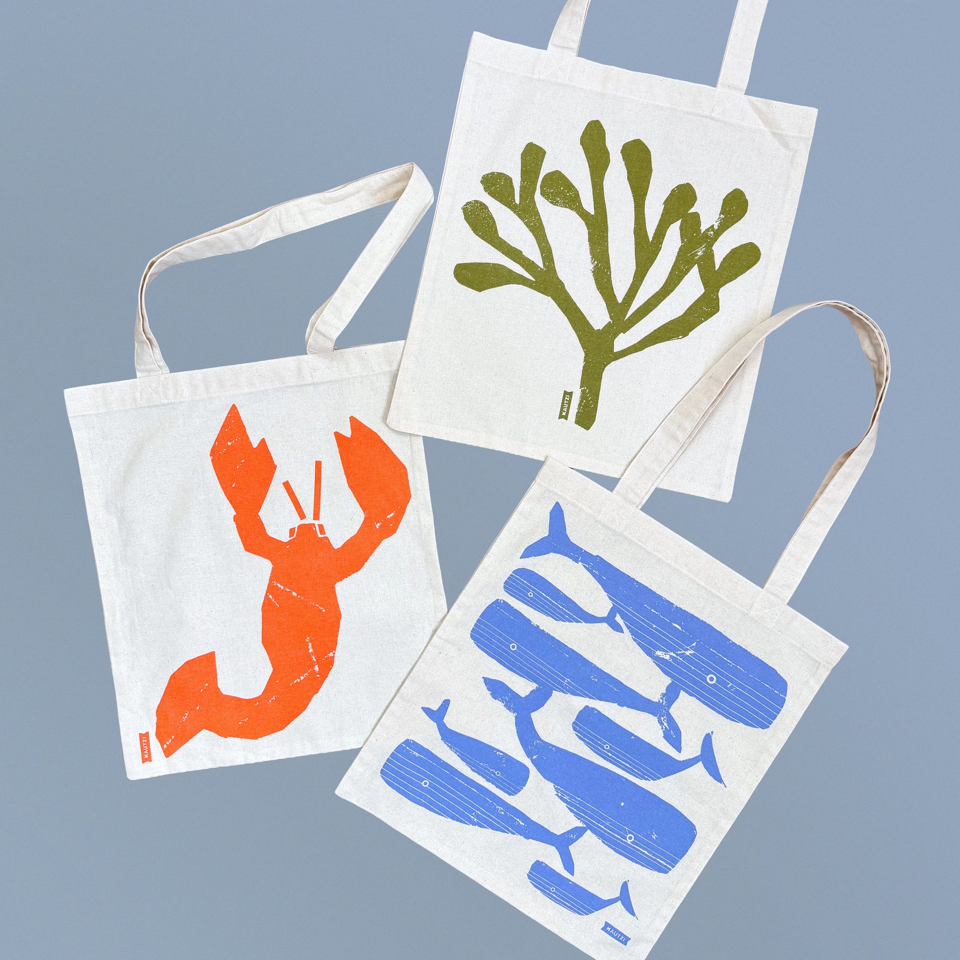 Kautzi tote bag collection featuring lobster, whale, and rockweed designs, shown together on blue background, Cape Breton, Nova Scotia, Canada.