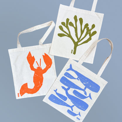 Kautzi tote bag collection featuring lobster, whale, and rockweed designs, shown together on blue background, Cape Breton, Nova Scotia, Canada.