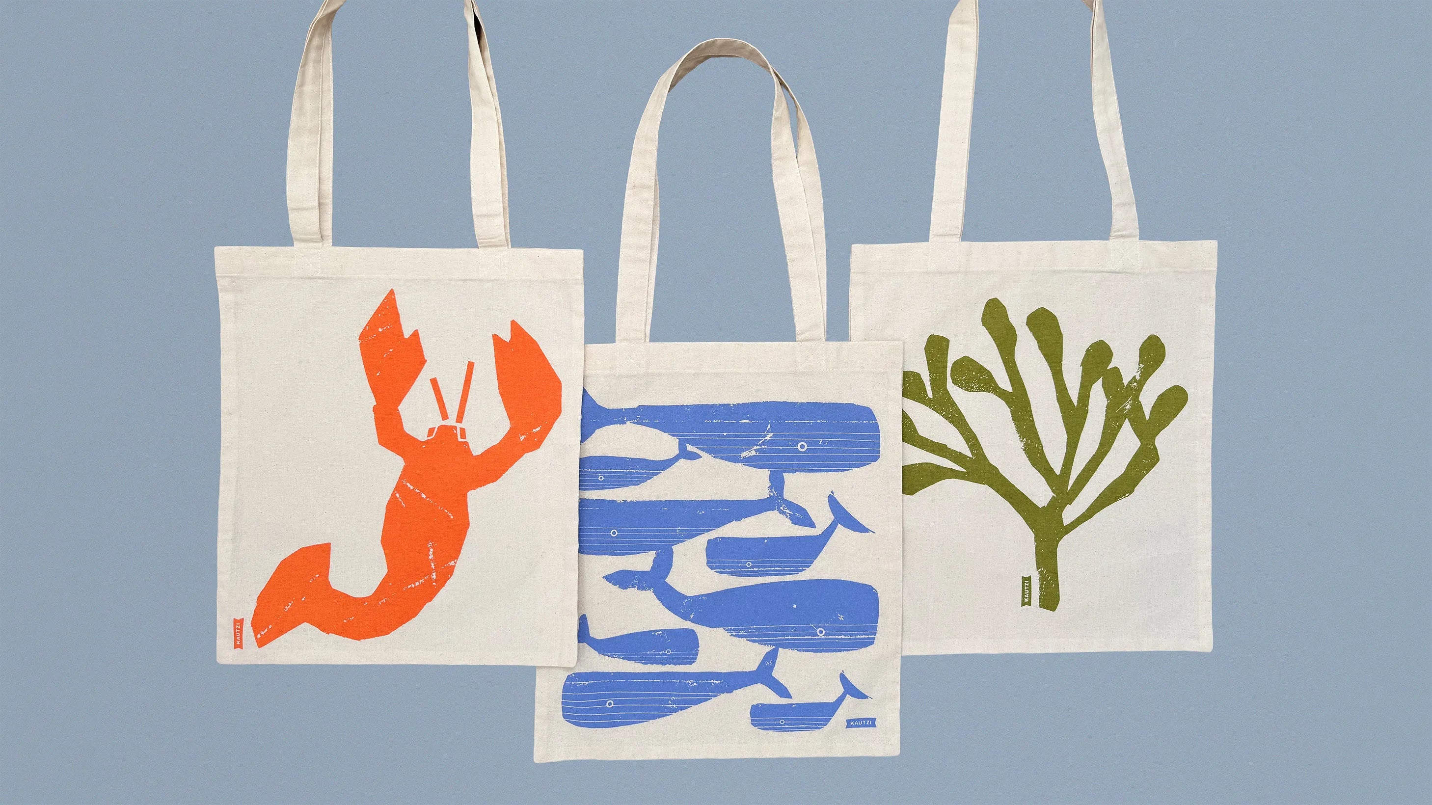 Three Kautzi illustrated cotton tote bags with East Coast sea life designs – lobster catch, whale pod, and rockweed – on a blue-grey background