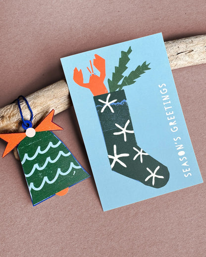 Kautzi sea bell wooden ornament from Cape Breton, Nova Scotia, shown beside a Season’s Greetings card with a lobster stocking illustration.