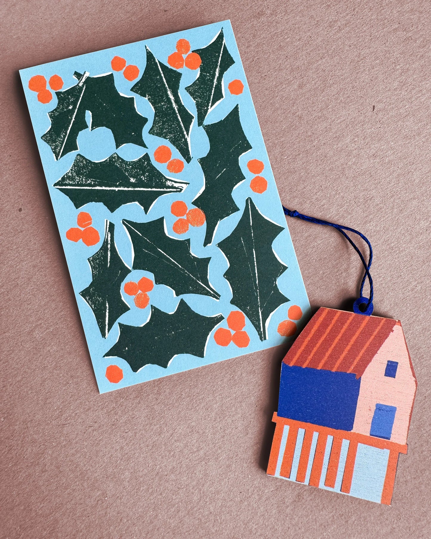 Kautzi fish shack wooden ornament from Cape Breton, Nova Scotia, shown next to a Christmas card with hollies and berries.