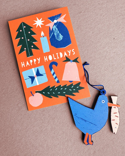 Kautzi seagull wooden ornament from Cape Breton, Nova Scotia, shown beside a Happy Holidays card in blue and orange tones.