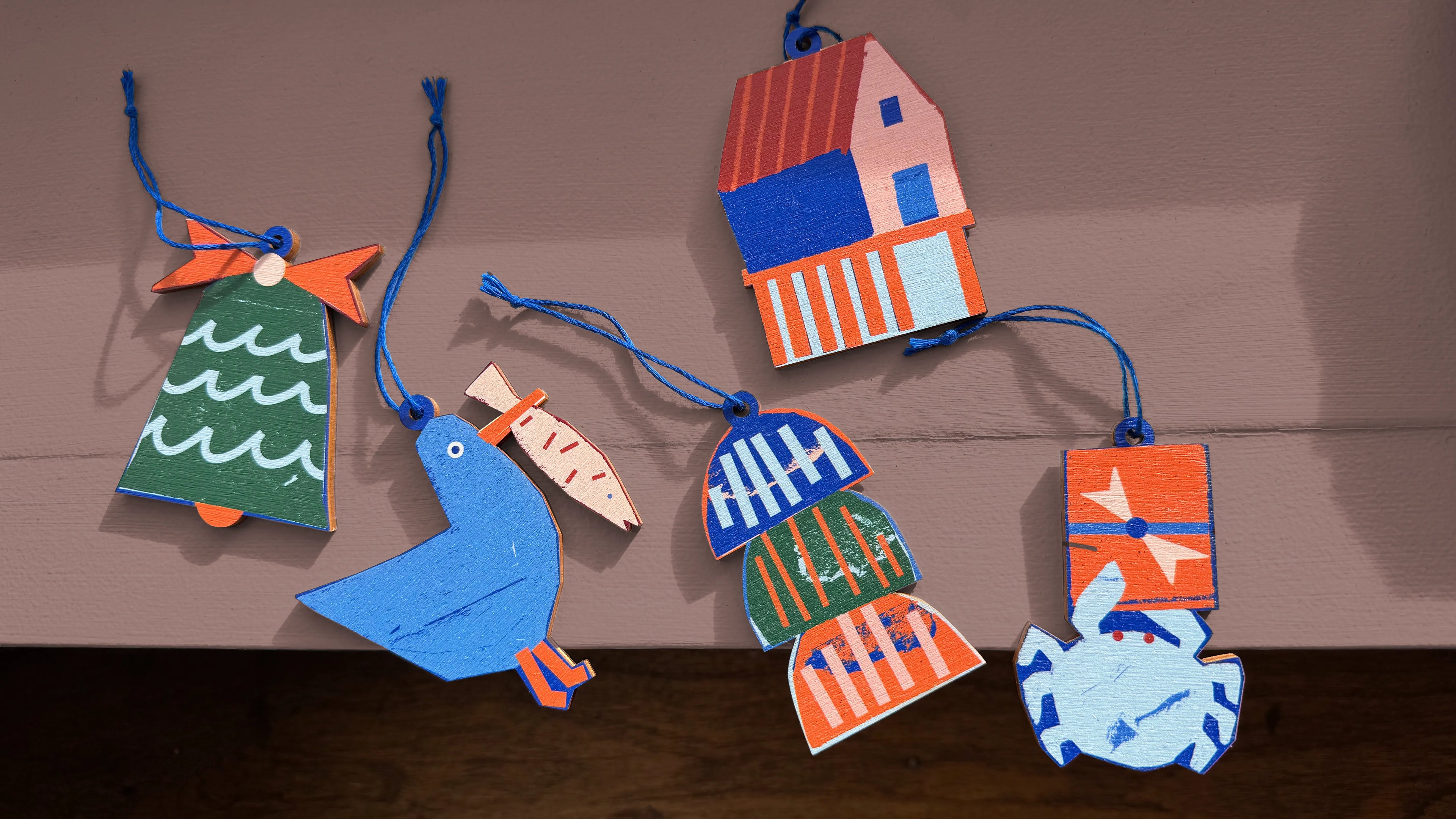 Kautzi wooden ornaments featuring five colourful East Coast designs – a bell, seagull, fish shack, lobster traps, and crab – displayed on a pink surface. Designed in Cape Breton, Nova Scotia, Canada.