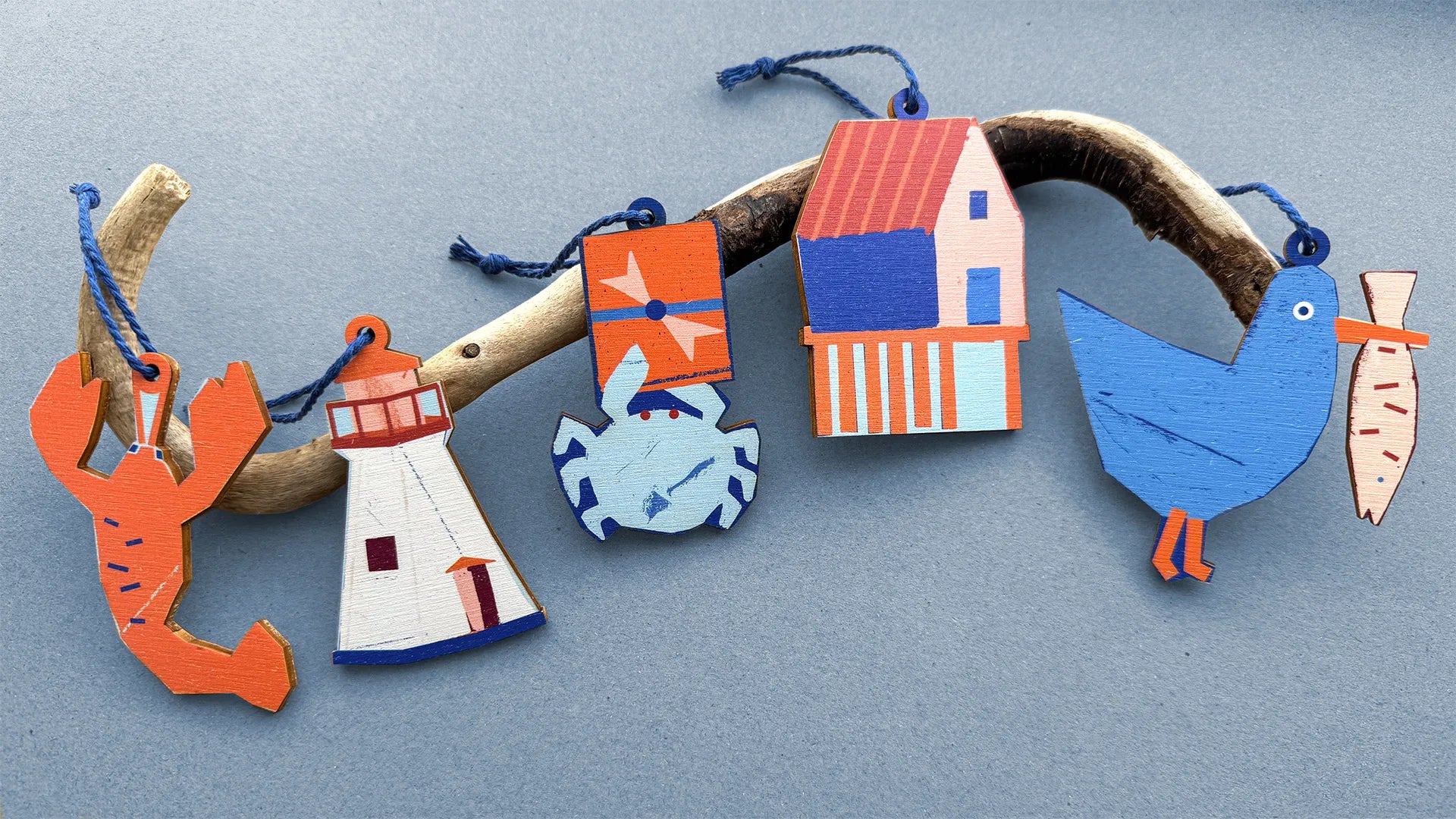 Five Kautzi wooden ornaments in Maritime motifs – lobster, lighthouse, crab, fishing shack, and seagull – hanging from a piece of driftwood