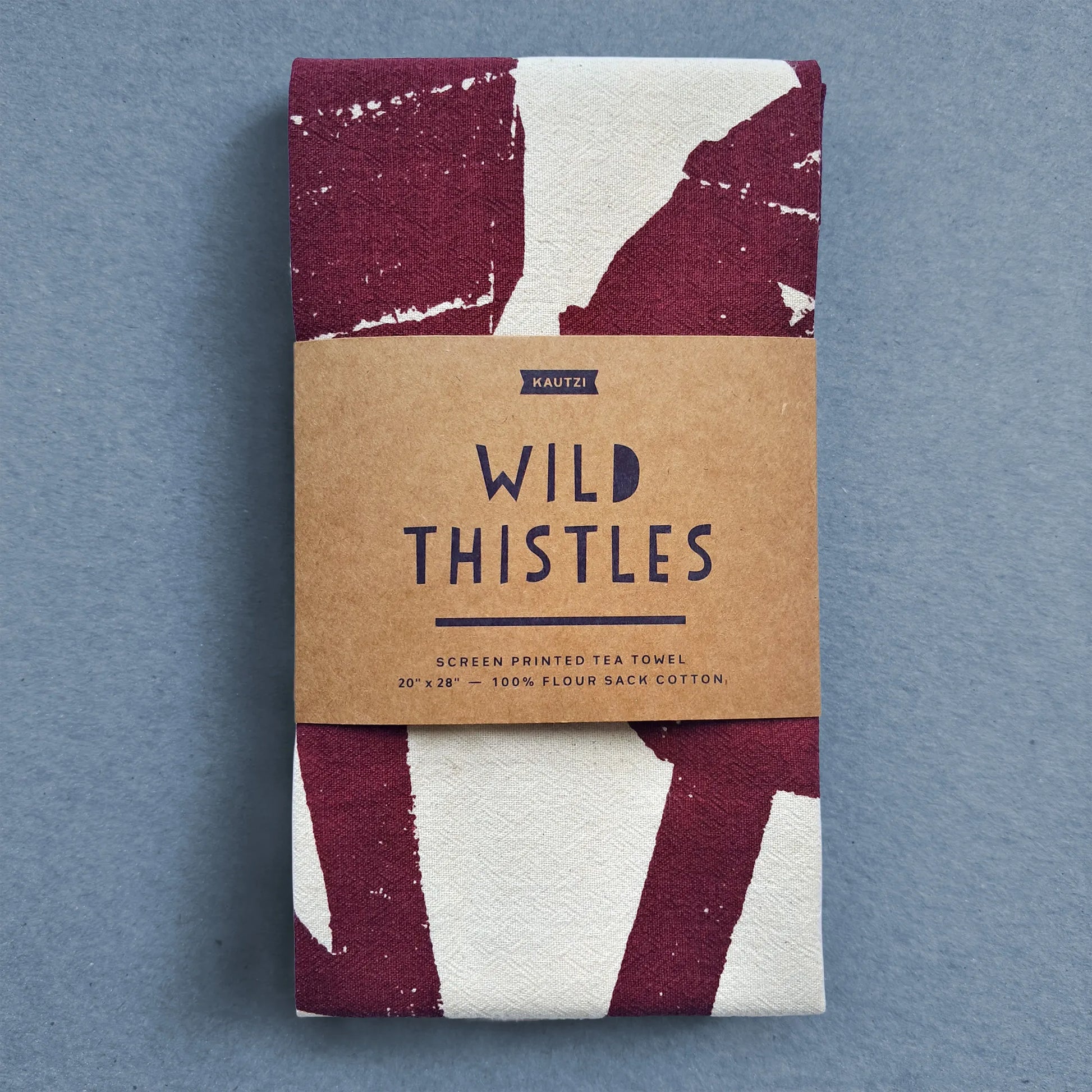 Kautzi Wild Thistles tea towel folded with kraft paper label, screen printed in deep plum on natural white cotton, photographed on blue background.