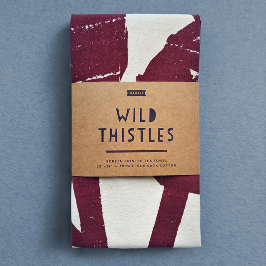 Kautzi Wild Thistles tea towel folded with kraft paper label, screen printed in deep plum on natural white cotton, photographed on blue background.